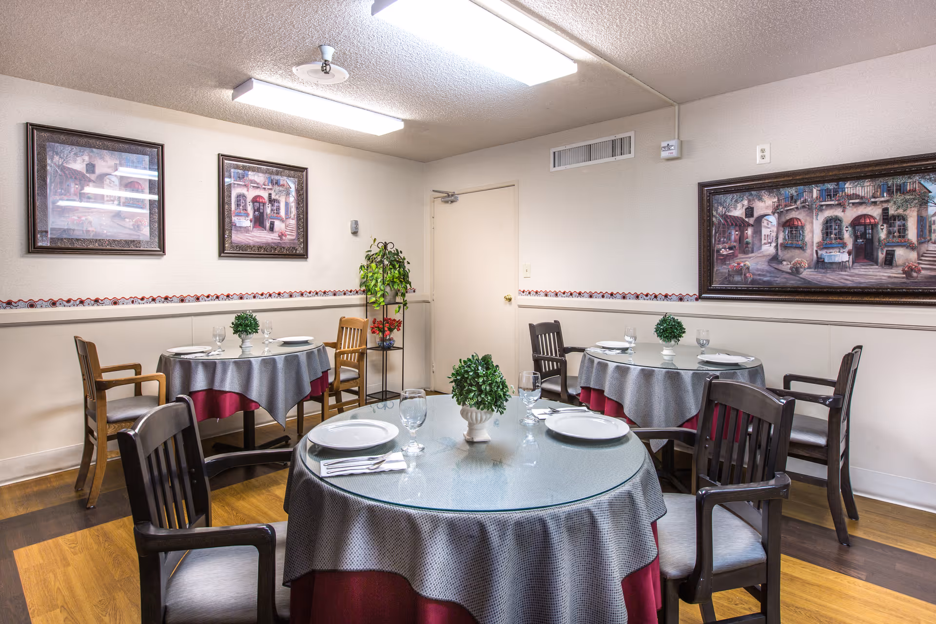 Small dining room with round tables set with plates and glasses, wooden chairs, framed paintings on the walls, and potted plants.