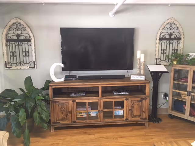 A living room area with a mounted flat-screen TV above a wooden media console. The console has shelves and cabinets with books and baskets inside. On top of the console are decorative items including a large letter 'C', candles, and a small sign. There are two decorative wrought iron wall hangings on either side of the TV. A large green plant is positioned to the left of the console, and a small stand with a book or menu is to the right. The floor is wooden.