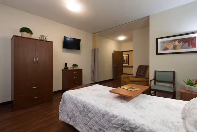 A senior living facility bedroom with a bed covered in a white quilt, a wooden wardrobe and dresser, a wall-mounted TV, an armchair, a wooden chair, a small table with a plant, and framed artwork on the wall.