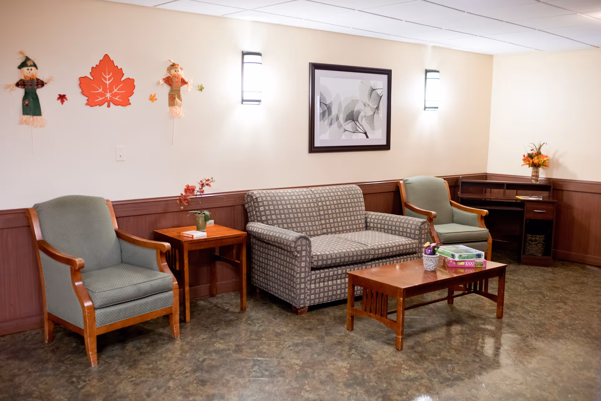 A cozy seating area in a senior living facility with two green armchairs, a patterned loveseat, a wooden coffee table with a puzzle box and a small flower pot, and a side table with a plant and books. The walls are decorated with autumn-themed decorations including two scarecrows and a large orange leaf, as well as a framed abstract artwork. The room has warm lighting and a wooden wainscoting along the walls.