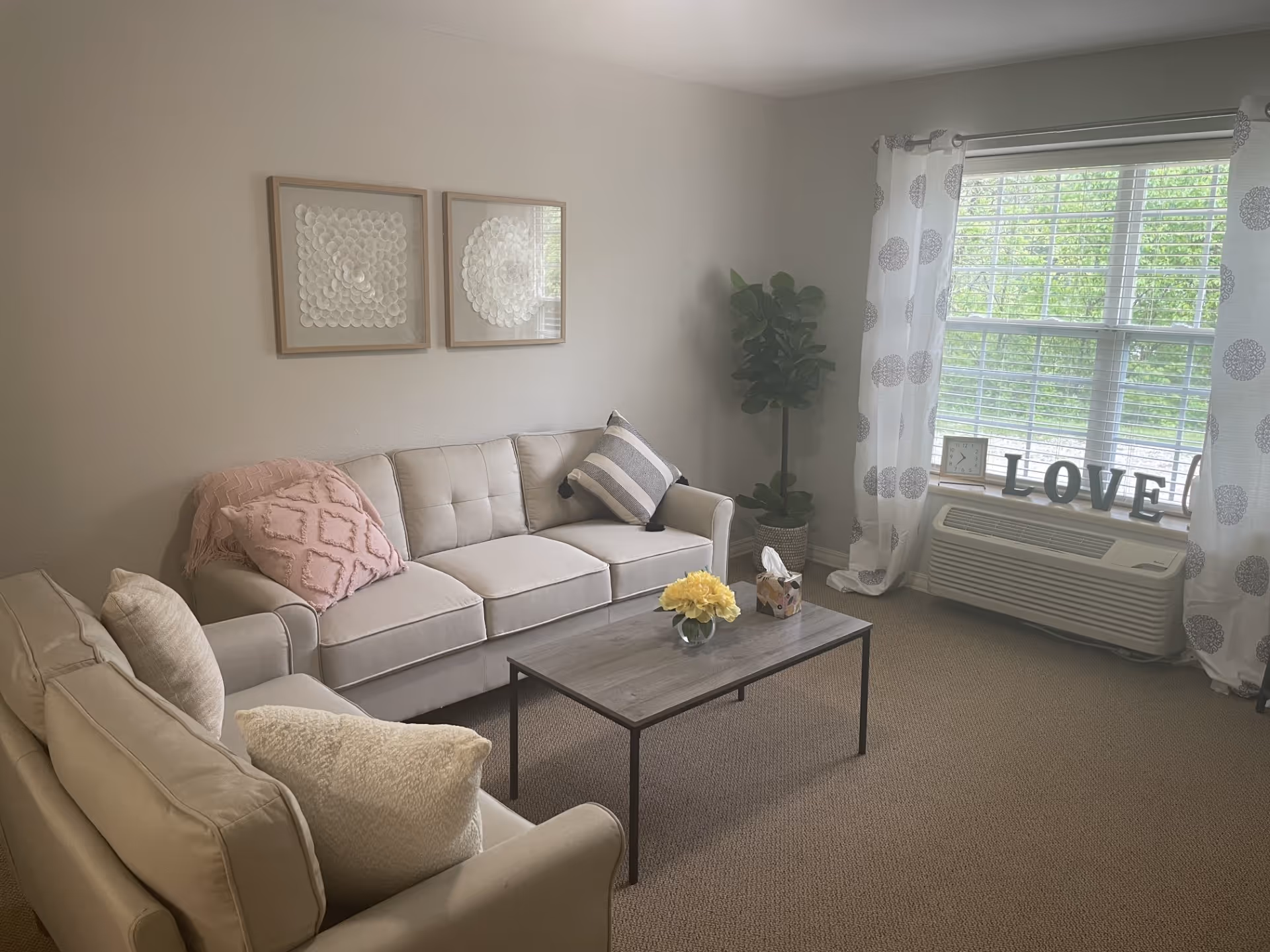 A cozy living room with two beige sofas adorned with cushions and a pink throw blanket. A wooden coffee table with a small vase of yellow flowers and a tissue box sits in the center. The room has a large window with white curtains featuring a circular pattern, and decorative letters spelling 'LOVE' on the windowsill. Two framed abstract artworks hang on the wall above one sofa, and a potted plant is placed in the corner near the window.