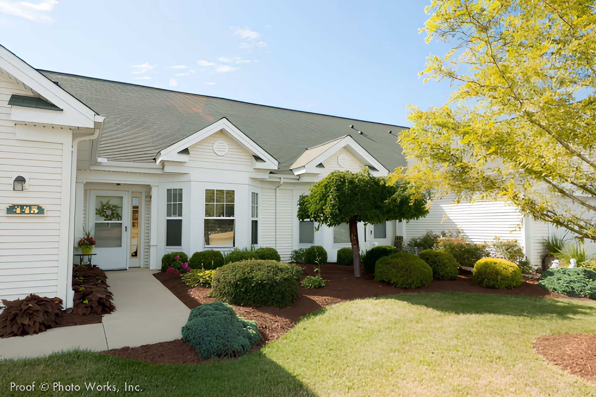 Exterior view of a white single-story building with a green roof, surrounded by well-maintained landscaping including bushes, small trees, and a grassy lawn under a clear blue sky.