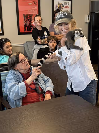 A group of people in a common area of a senior living facility. An elderly woman seated at a table is touching the tail of a lemur held by a smiling woman wearing a cap and white shirt. Three other people, including a young boy and two adults, watch the interaction in the background. Posters are visible on the wall behind them.