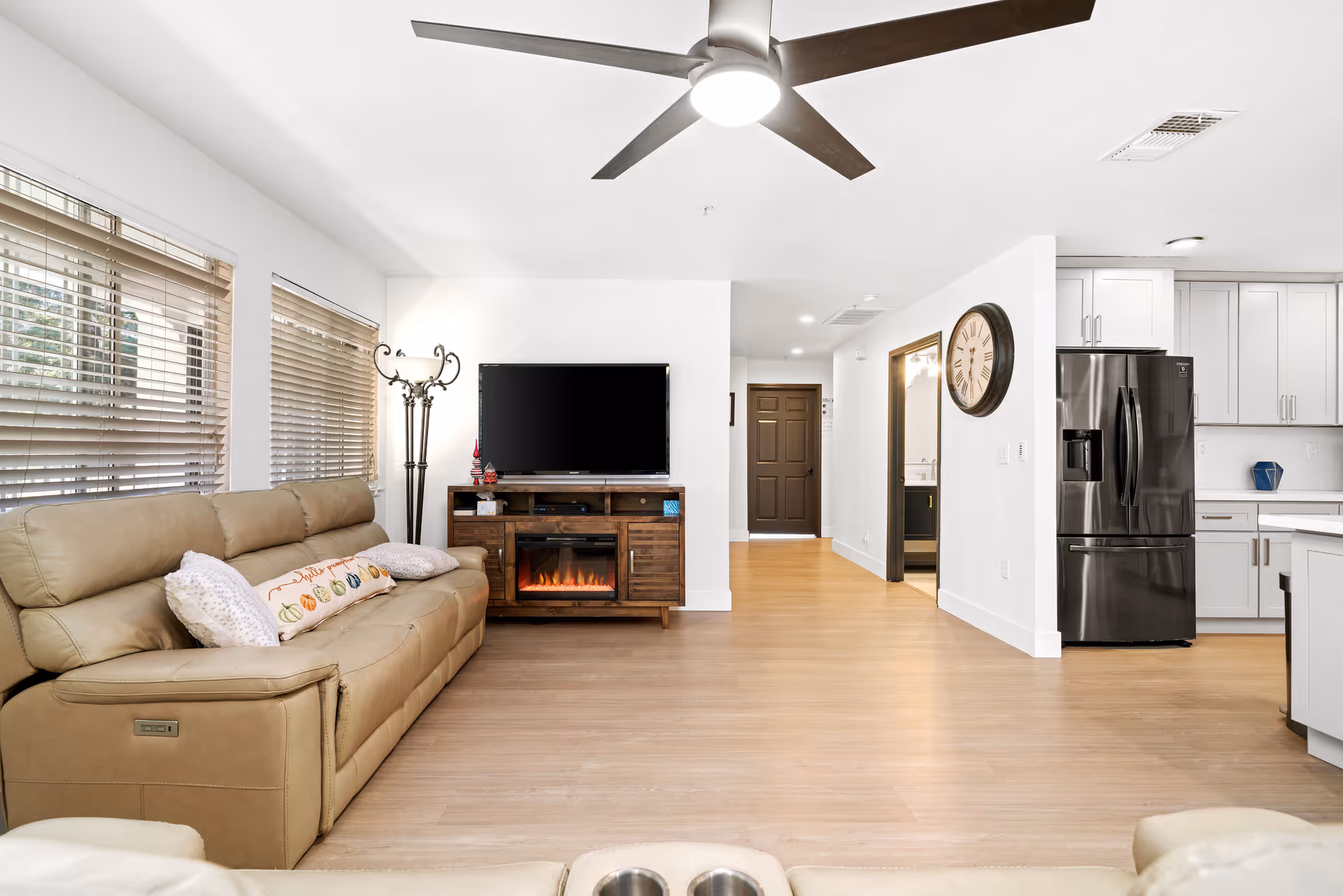 A bright and modern living room area with a beige leather sofa adorned with decorative pillows, large windows with blinds, a ceiling fan, a flat-screen TV mounted on a wooden stand with an electric fireplace below, and a view into a kitchen with white cabinets and a black refrigerator.