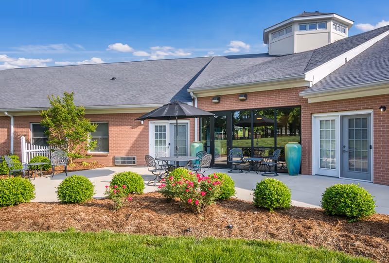 Outdoor patio area of a brick building with a gray roof, featuring several metal tables and chairs, one table with a black umbrella. The patio is surrounded by neatly trimmed bushes and flowering plants, with a well-maintained lawn in the foreground under a blue sky with some clouds.