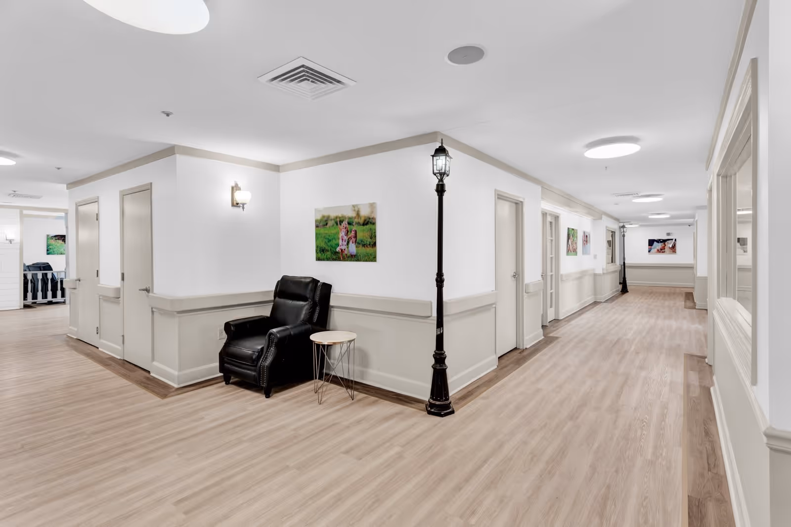 A clean, well-lit hallway in an assisted living facility with light wood flooring and white walls. The hallway features several closed doors, a black leather armchair with a small round side table, decorative streetlamp-style light fixtures, and framed photos on the walls.