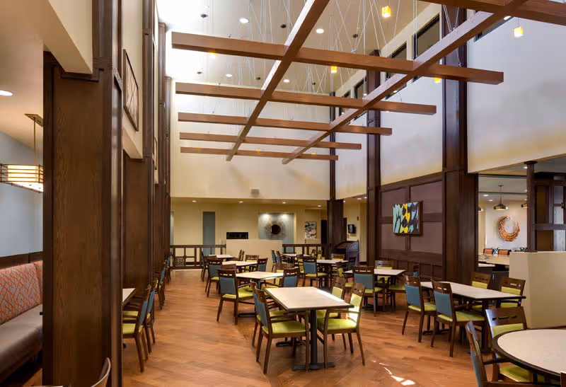 A spacious dining room with wooden floors and high ceilings featuring exposed wooden beams. The room is furnished with multiple tables and chairs, some with green cushions and others with blue cushions. There is a long bench with orange patterned upholstery along one wall. The walls are decorated with modern artwork, and the room is well-lit with natural and artificial lighting.