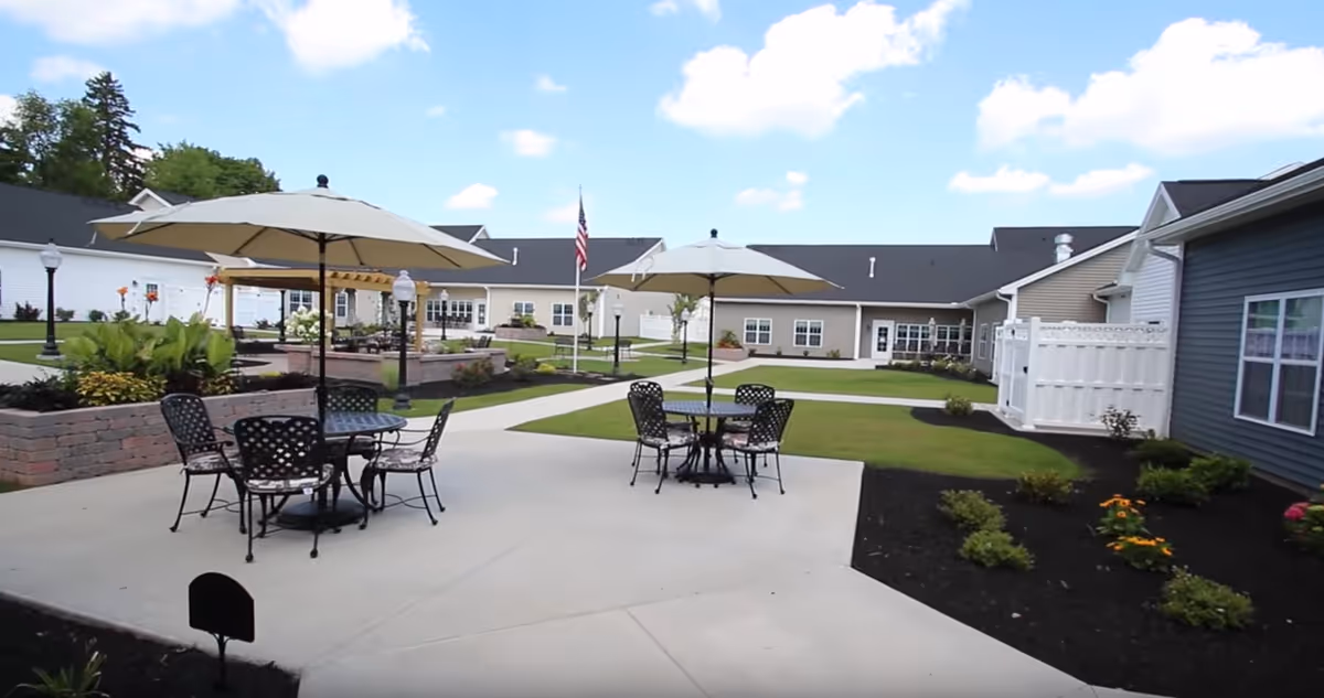 Outdoor courtyard with patio tables and umbrellas, landscaped plantings, and surrounding single-story senior living buildings under a blue sky.