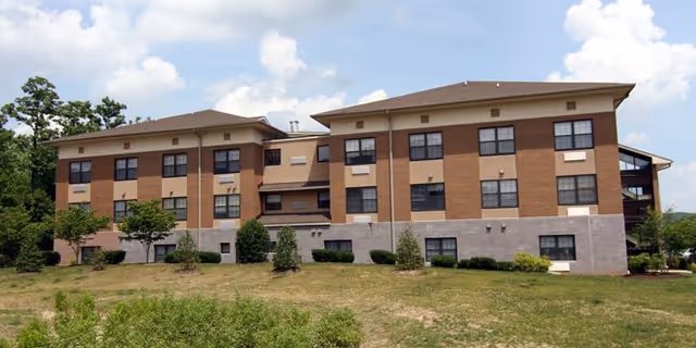 Exterior view of a three-story senior living facility building with a beige and brown facade, multiple windows, and a grassy area with shrubs in front under a partly cloudy sky.