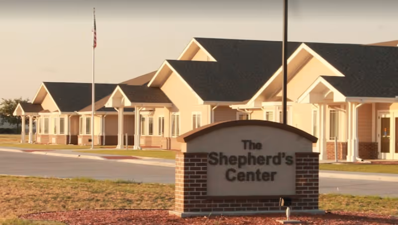 Exterior view of The Shepherd's Center building with a sign in the foreground displaying the facility's name. The building has a beige exterior with white trim and multiple peaked roofs. There is a flagpole with an American flag near the building.