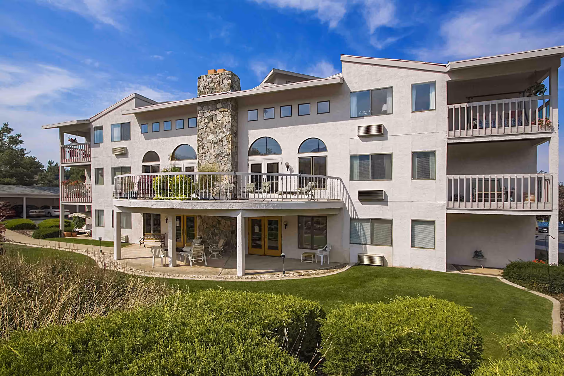 Three-story white retirement community building with balconies, a central stone chimney, and a manicured lawn.