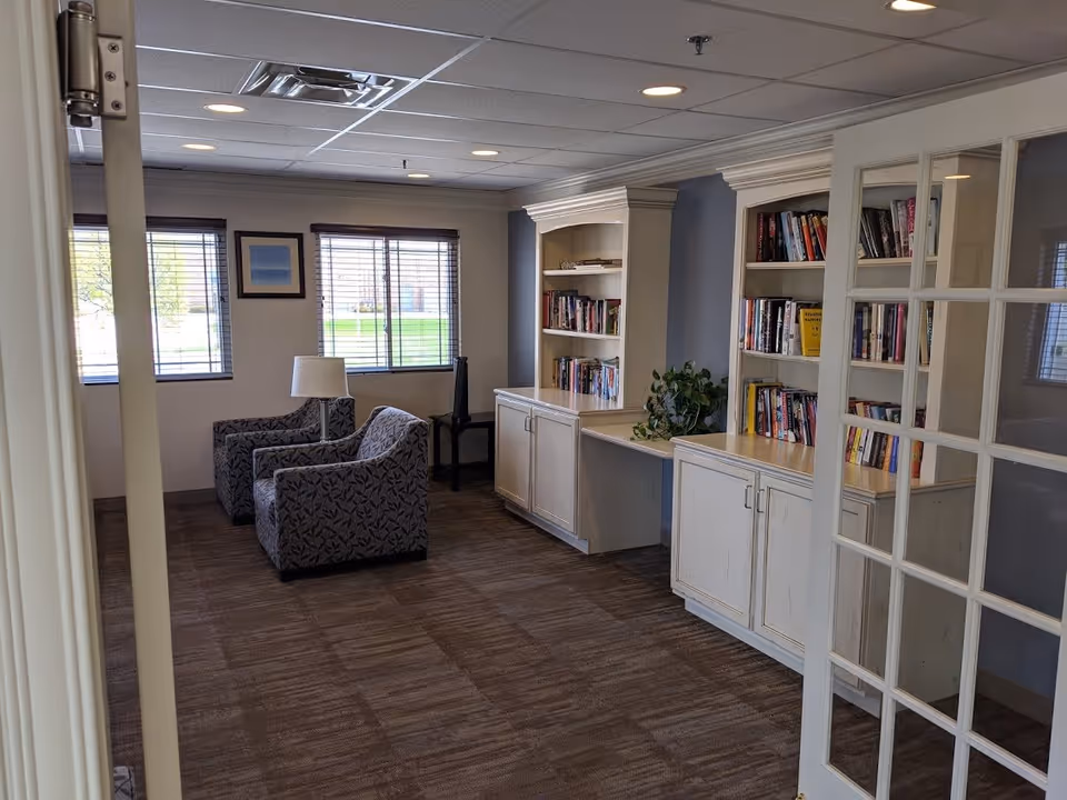 A cozy sitting area in Franciscan Court Supportive Living with two patterned armchairs, a small table with a lamp between them, large windows with blinds, and built-in white bookshelves filled with books and a potted plant. The room has carpeted flooring and a ceiling with recessed lighting.