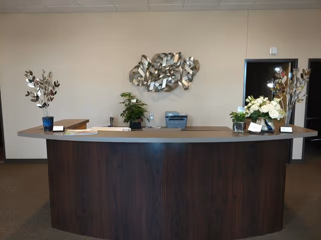 A reception desk with a dark wood front and a light-colored countertop. On the desk are decorative plants and flowers in vases, along with some papers and office supplies. Behind the desk is a beige wall with a modern metallic wall sculpture and a printer. There is an open doorway to the right.
