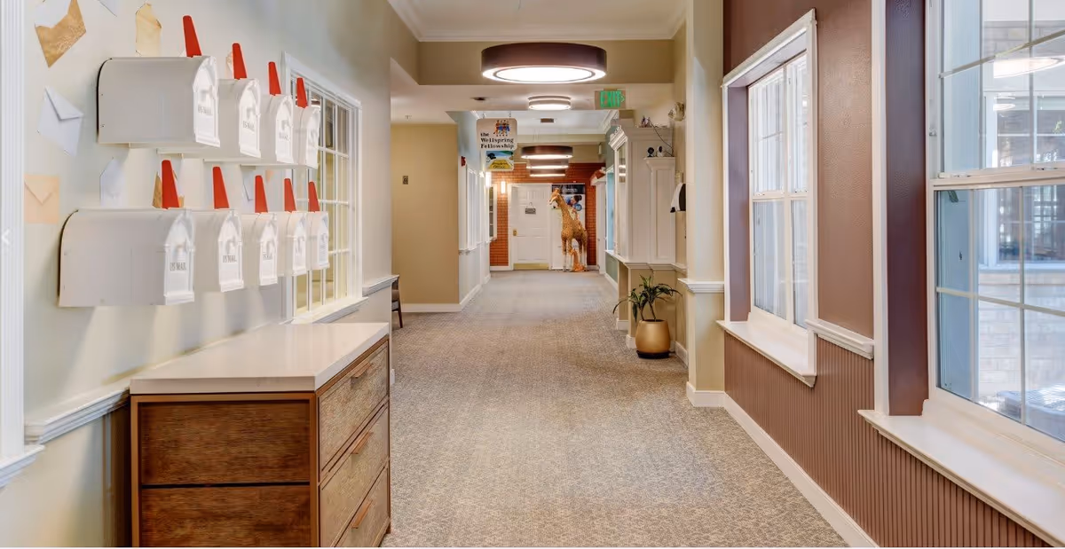 A well-lit hallway in a senior living facility with multiple white mailboxes mounted on the left wall above a wooden cabinet. The hallway has beige walls, carpeted floor, and windows on the right side letting in natural light. At the end of the hallway, there is a door with a giraffe decoration and a sign hanging from the ceiling.