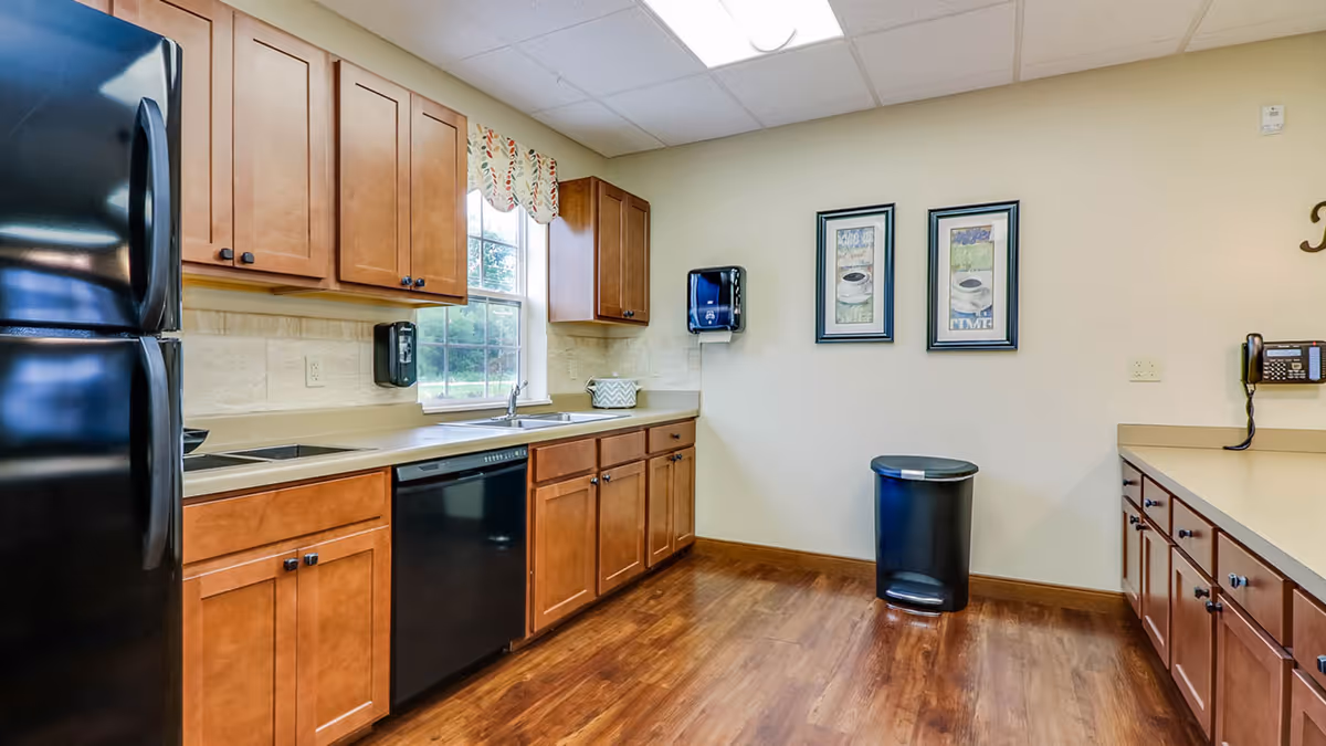 A clean kitchen area with wooden cabinets, a black refrigerator, dishwasher, and sink under a window with a patterned valance. The kitchen has a light beige countertop and wood flooring. On the wall, there are two framed pictures and a mounted paper towel dispenser. A black trash can is placed against the wall, and a telephone is mounted on the right side.