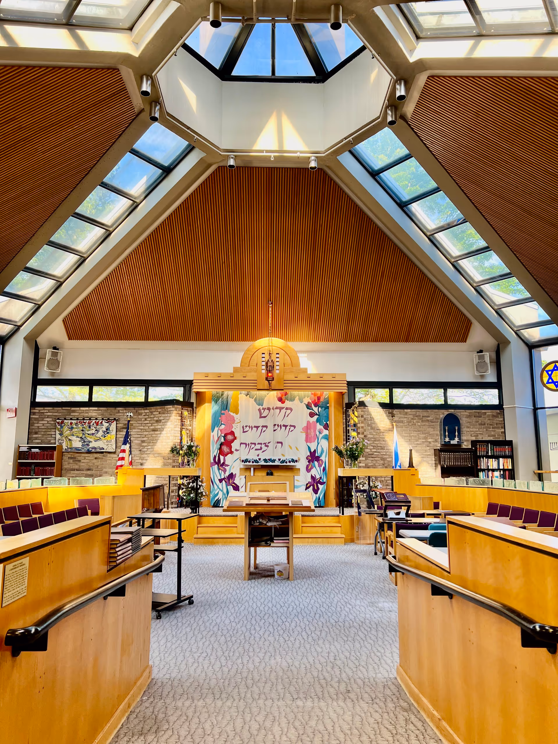 Bright, modern sanctuary interior with a central bimah and ark beneath a skylit vaulted wooden ceiling, flanked by pews and a decorative Hebrew tapestry.