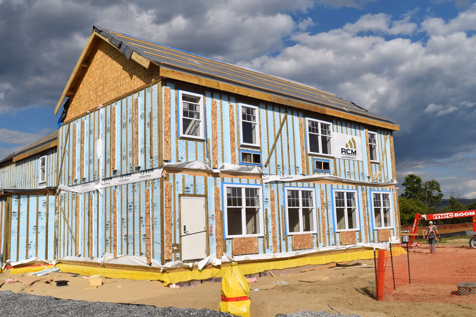 A two-story building under construction with exposed wooden framing and blue insulation panels. Several windows are installed, and the roof is partially covered. A construction worker and equipment are visible on the right side, with a partly cloudy sky in the background.