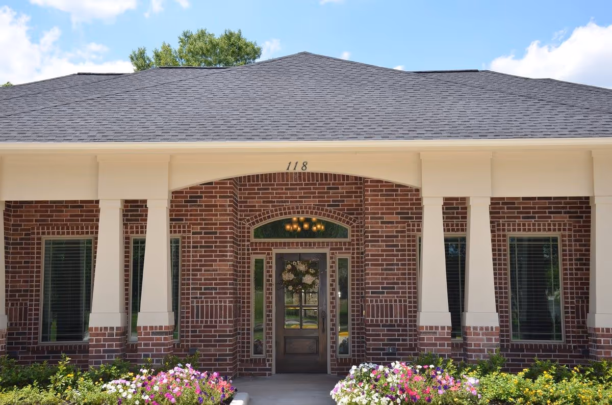 Front exterior view of a brick building with a covered entrance supported by white columns, a wooden door with glass panels, and flower beds with colorful flowers in front.
