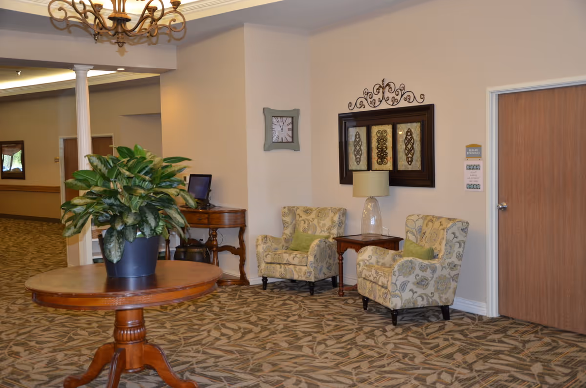 Interior view of a nursing facility hallway with a round wooden table holding a large potted plant in the foreground. Two patterned armchairs with green cushions are placed beside a small wooden side table with a lamp. Wall decorations include a clock and framed artwork. The floor is carpeted with a leaf pattern, and a chandelier hangs from the ceiling.