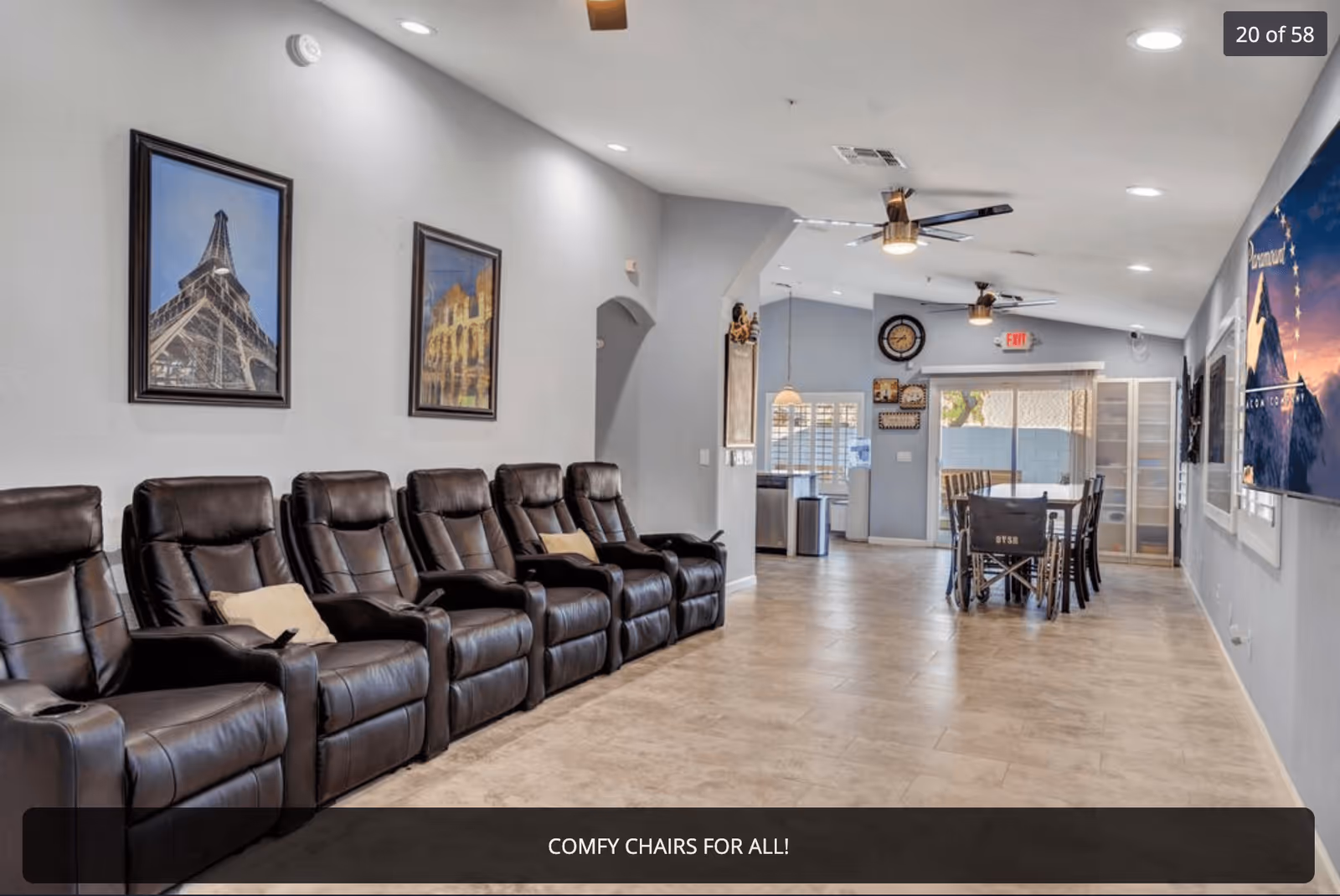 A spacious living area with a row of six black leather recliner chairs along the left wall, each with a small pillow. The walls are painted light gray and decorated with framed pictures. The room has a tiled floor and ceiling fans with lights. In the background, there is a dining table with chairs and a wheelchair, near a glass sliding door leading outside. A large TV is mounted on the right wall.