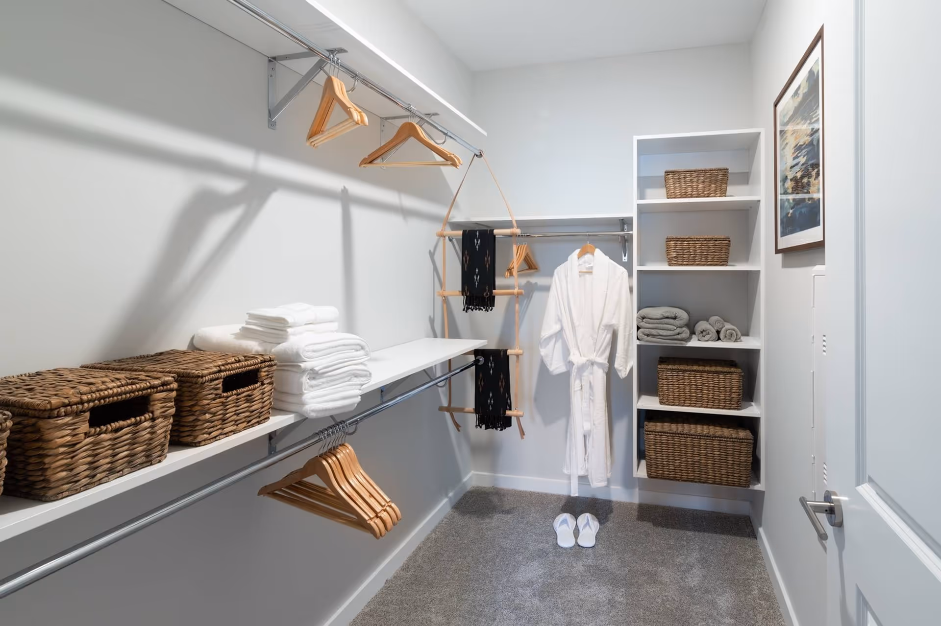 A bright organized walk-in closet with shelves holding wicker baskets and folded towels, wooden hangers, a white robe and slippers on the carpeted floor.