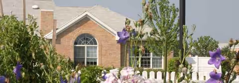 Luther Acres exterior: brick building with arched window, white fence, purple flowers.
