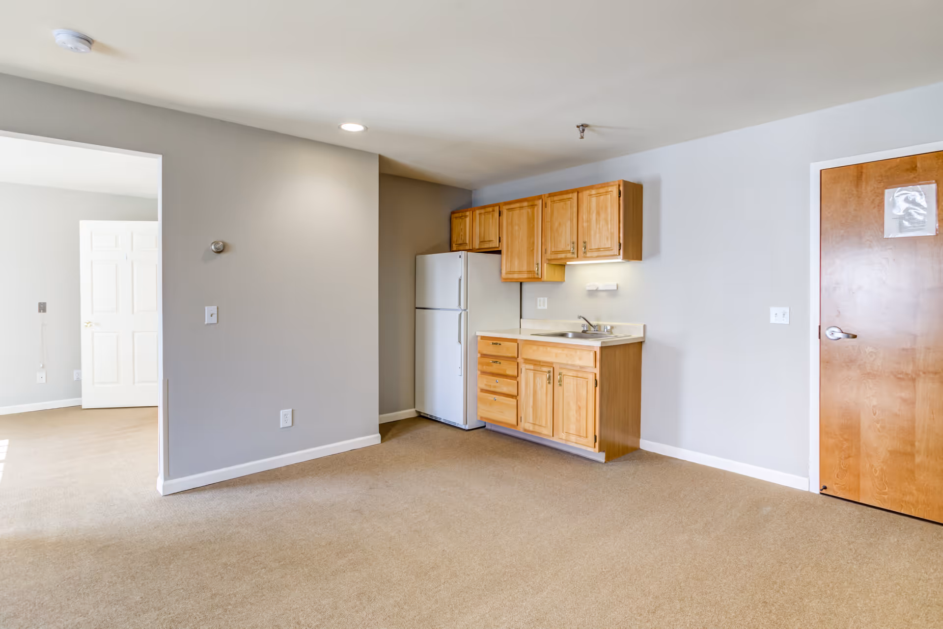 An interior view of a small kitchen area in an assisted living facility. The kitchen features light wooden cabinets, a white refrigerator, a countertop with a sink, and beige carpeted flooring. To the right is a wooden door with a paper attached, and to the left is an open doorway leading to another room with white double doors.