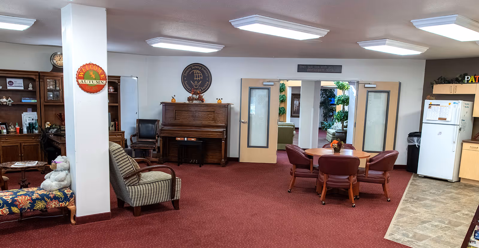 A cozy common area in a senior living facility featuring a piano against the wall, a round table with four chairs, a striped armchair, and a wooden cabinet with various items. The room has red carpet flooring and bright ceiling lights. There is a white refrigerator and kitchen cabinets on the right side, and double doors leading to another room with plants visible inside. A sign on the wall above the piano reads 'Seniors Serving Seniors'.