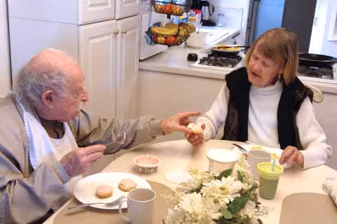 Two elderly adults sit at a dining table in a kitchen area sharing snacks and drinks.