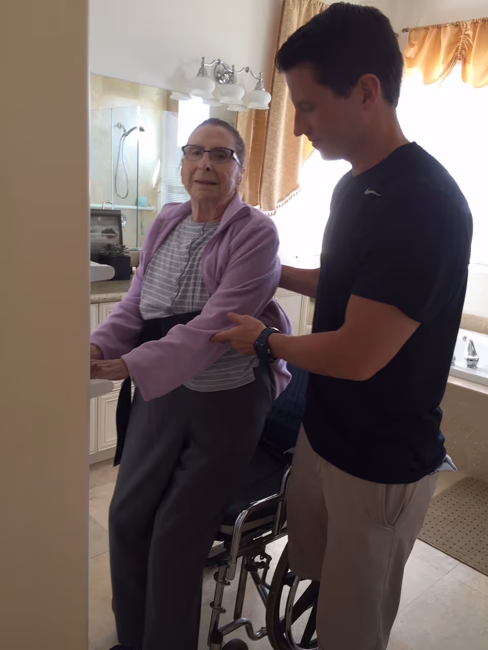 An elderly woman wearing glasses and a purple cardigan is being assisted by a man in a black shirt and beige pants as she stands next to a wheelchair in a bathroom. The bathroom has a shower with glass doors, a sink, and a bathtub with a window covered by a curtain.