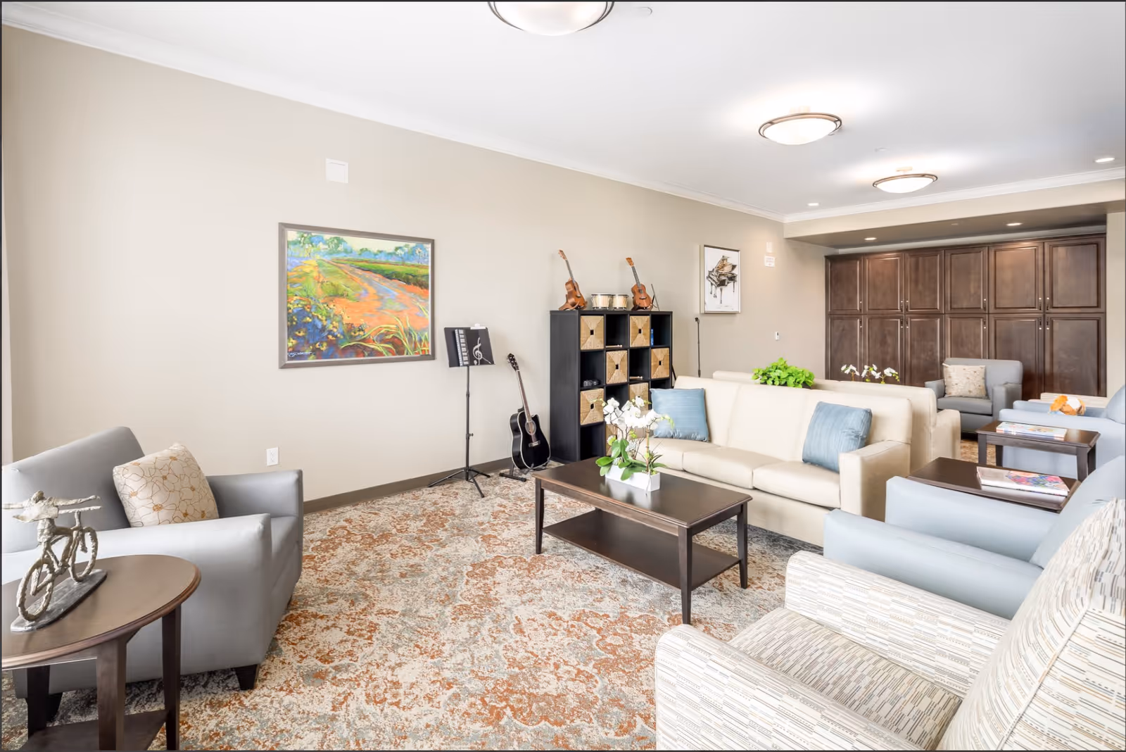 A bright and spacious living room with beige walls and a patterned carpet. The room features a cream-colored sofa with blue pillows, several armchairs in gray and patterned fabric, dark wood coffee tables, and a black shelving unit holding musical instruments including guitars and violins. There are framed paintings on the walls and built-in dark wood cabinets in the background.