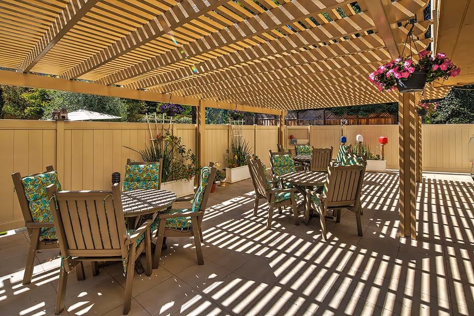 Outdoor patio area with wooden pergola casting striped shadows on the floor. Several round tables with wooden chairs featuring green floral cushions are arranged under the pergola. Hanging flower pots with pink flowers are attached to the pergola beams. The area is enclosed by a beige fence with plants and greenery visible in the background.
