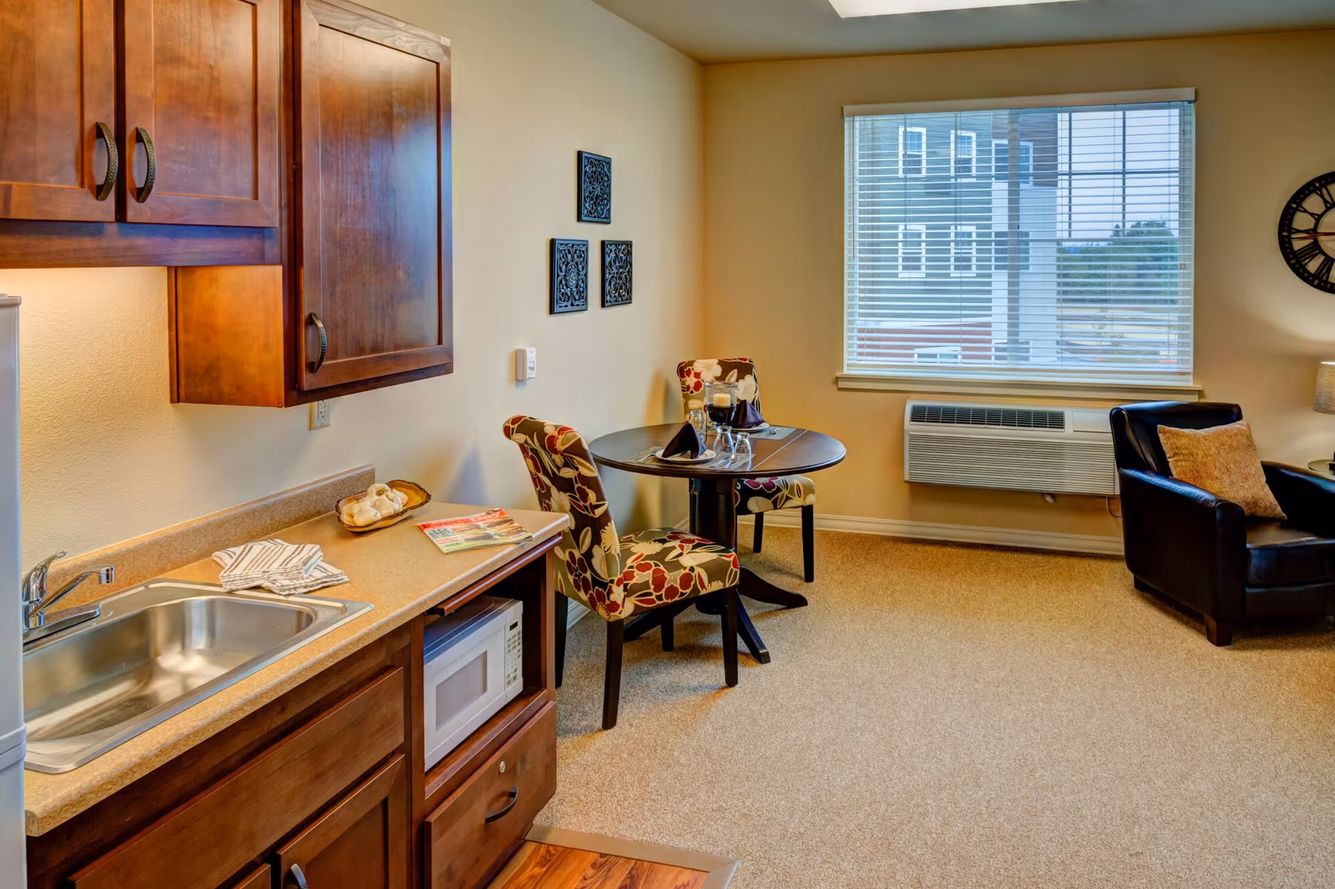 A cozy assisted living apartment interior featuring a small kitchenette with a sink, wooden cabinets, and a microwave. Adjacent to the kitchenette is a round dining table with two floral-patterned chairs. A window with blinds lets in natural light, and a black leather armchair with a beige cushion is positioned near the window. The walls are decorated with three small black wall hangings and a large clock.