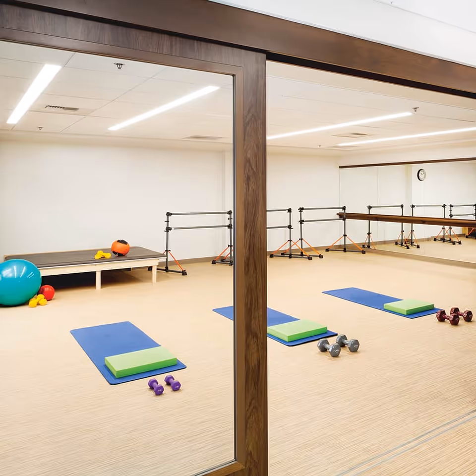 Exercise room with wooden floor and mirrored wall, featuring blue exercise mats each with a green cushion and pairs of dumbbells. There are also large exercise balls and small weights on the floor, and ballet barres along the mirrored wall.