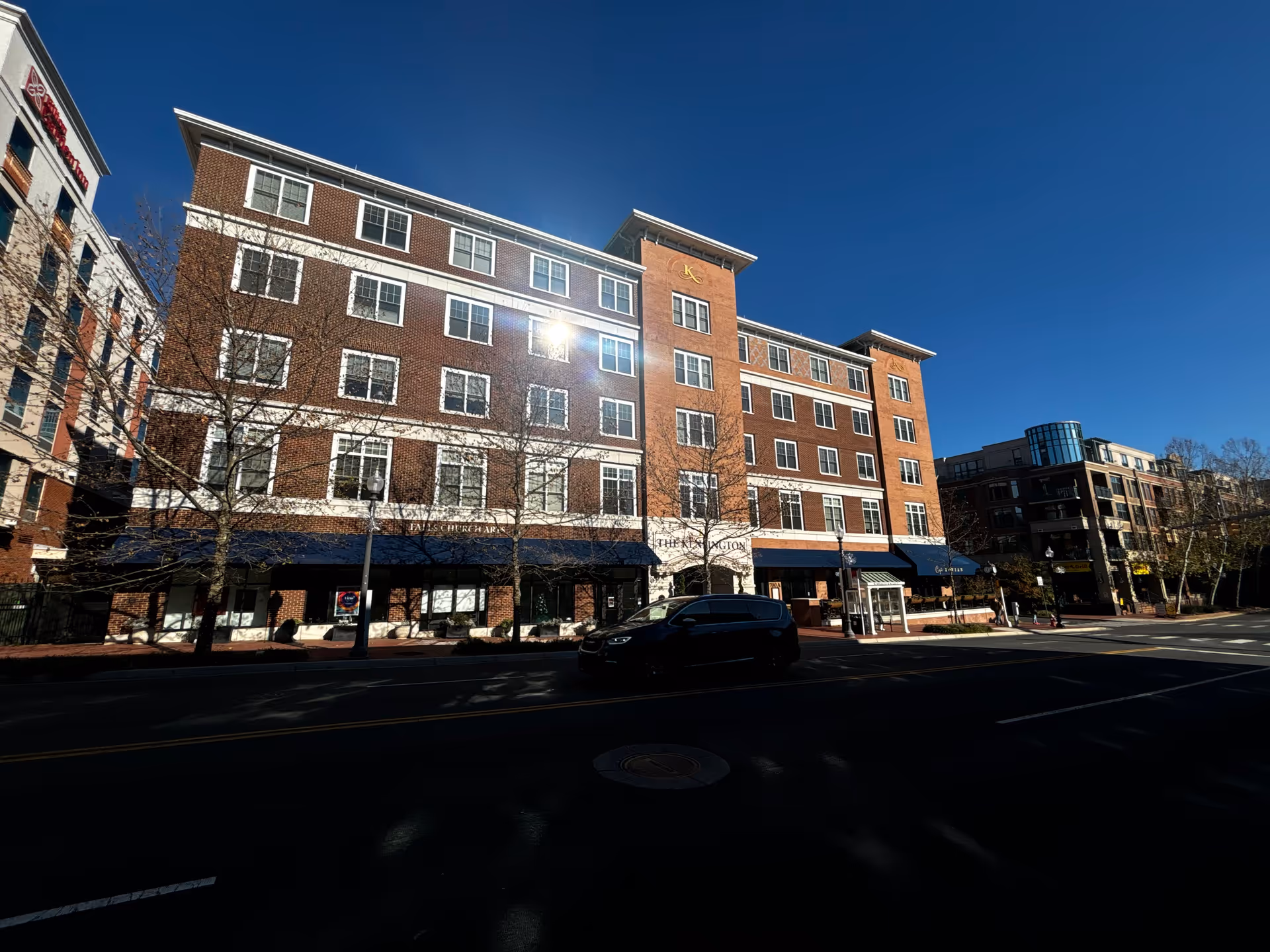 Street-level view of a five-story brick building with blue awnings, storefronts and a car parked on the road under a clear blue sky.