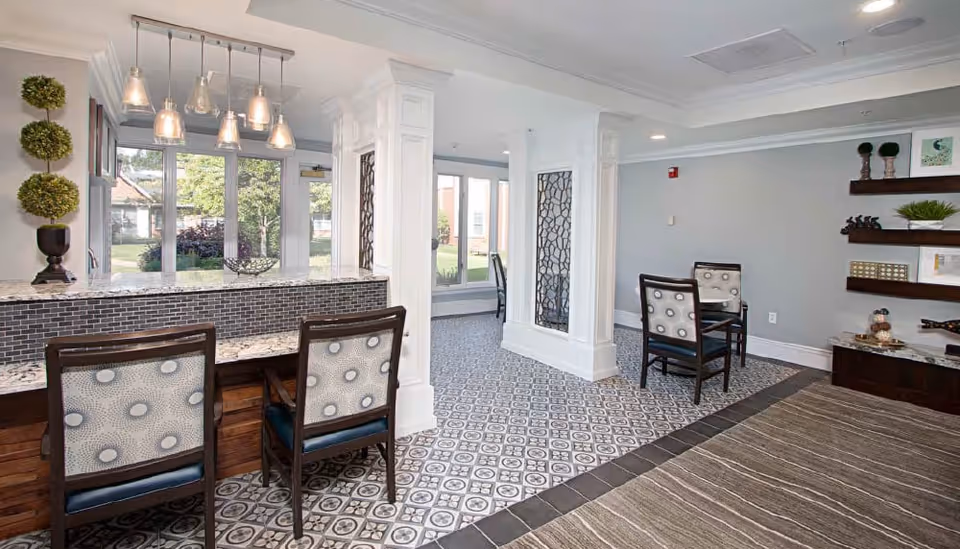 A bright and spacious interior common area with patterned tile flooring and a carpeted section. There are two chairs with patterned upholstery at a granite-topped counter with pendant lights hanging above. The room features white columns with decorative panels, large windows letting in natural light, and a seating area with two chairs and a small table. Shelves on the wall hold decorative items and plants.