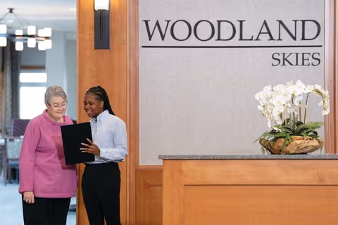 An elderly woman in a pink sweater is standing next to a younger woman in a white shirt and black pants who is holding and showing a black folder. They are inside a senior living community near a reception desk with a sign on the wall that reads 'WOODLAND SKIES'. There is a decorative plant with white flowers on the desk.