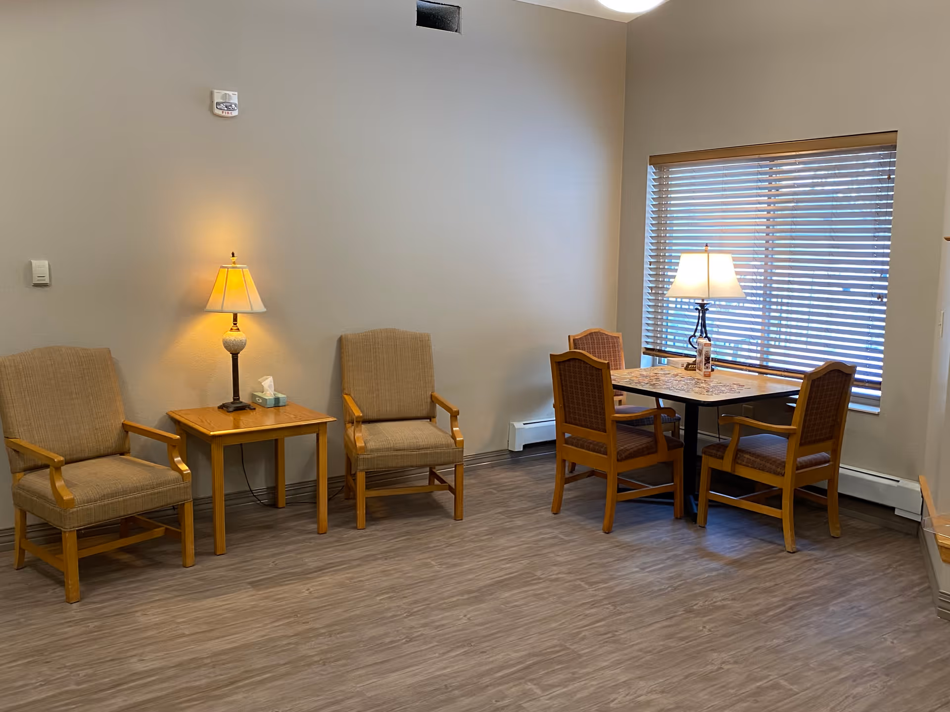 A cozy seating area in a senior community with two beige armchairs and a wooden side table with a lamp and tissue box. Nearby, there is a square table with four wooden chairs, a lamp on the table, and a window with blinds letting in natural light.