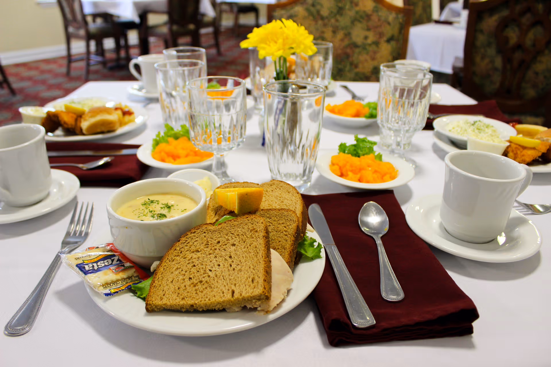 A dining table set with plates of food including sandwiches with rye bread, a bowl of creamy soup, a lemon wedge, and a packet of crackers. There are also small bowls of mashed sweet potatoes garnished with parsley, cups and glasses, silverware on burgundy napkins, and a yellow flower centerpiece. The background shows more tables and chairs in a dining room setting.