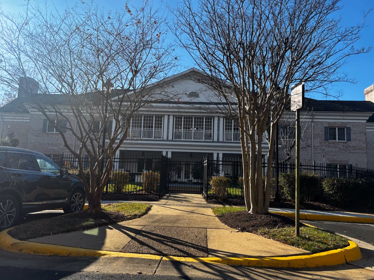 Front exterior of a two-story brick senior living building with a gated entrance, walkway, parked car, and leafless trees.