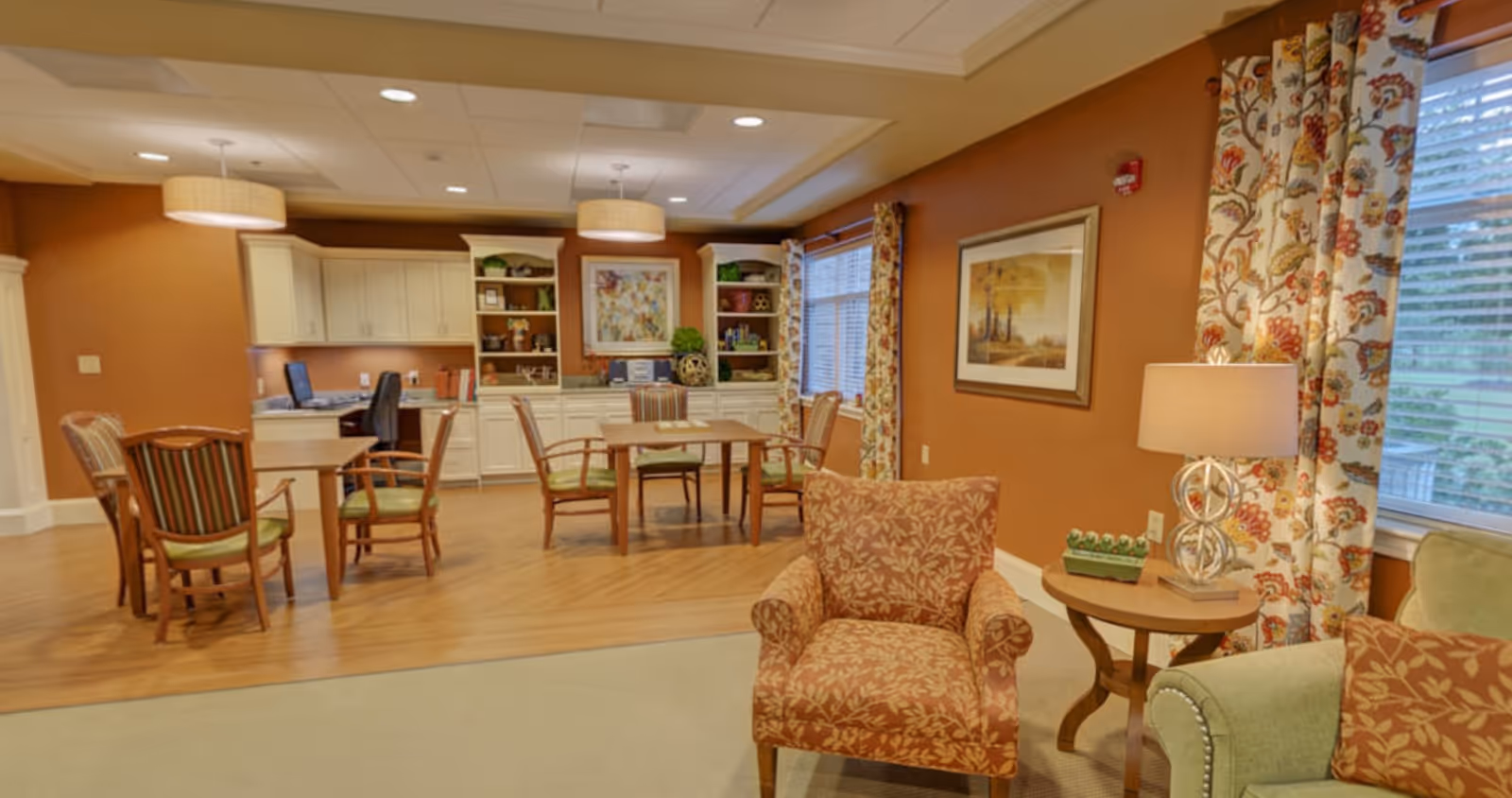 Bright communal lounge and dining area with tables, chairs, built-in cabinetry and upholstered armchairs.