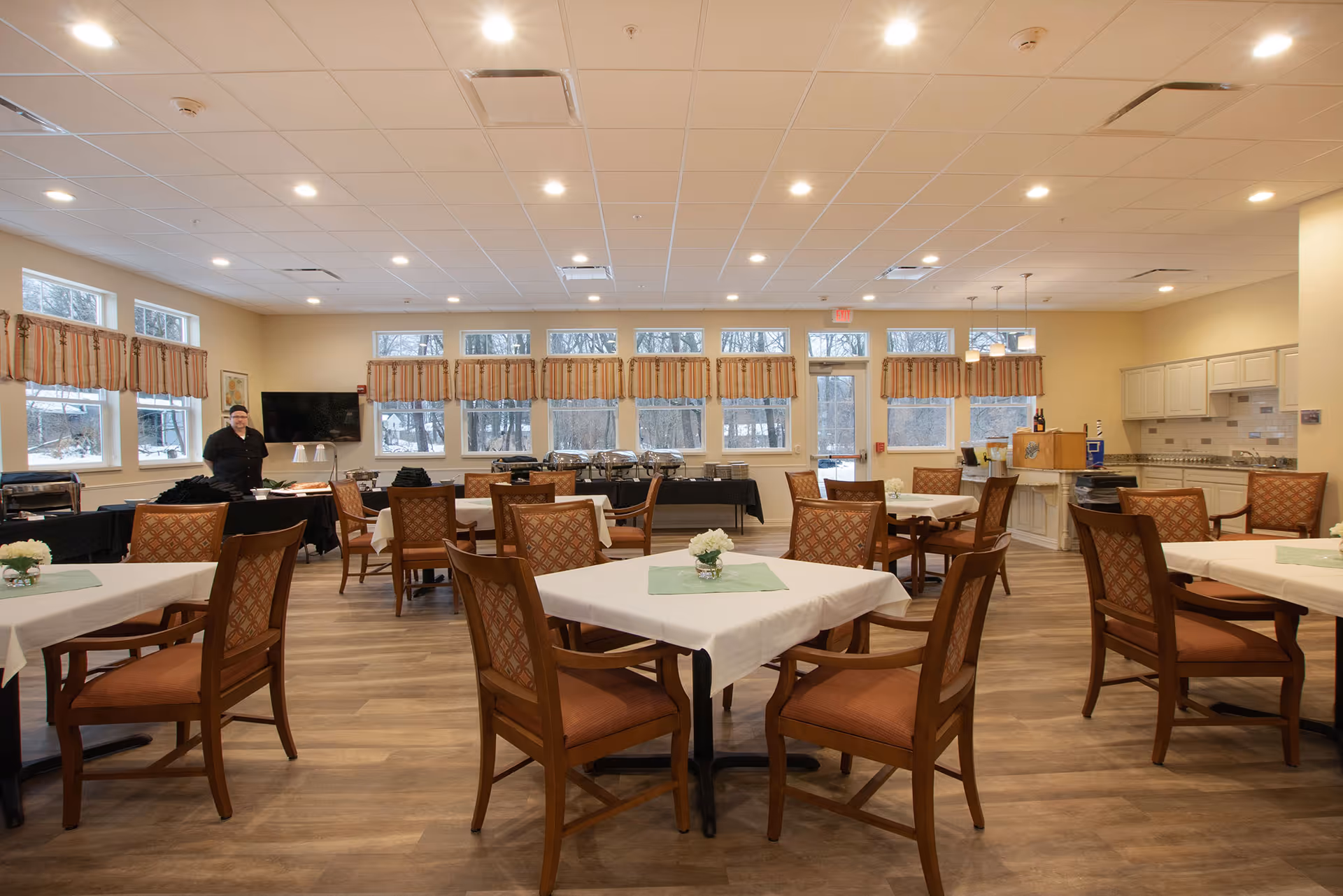 A spacious dining room with multiple tables covered in white tablecloths and green placemats, each surrounded by wooden chairs with cushioned seats. The room has large windows with striped valances, allowing natural light to enter. A person stands near a table on the left side of the room. The back wall features a buffet setup with chafing dishes and a kitchen area with cabinets and countertops on the right side.