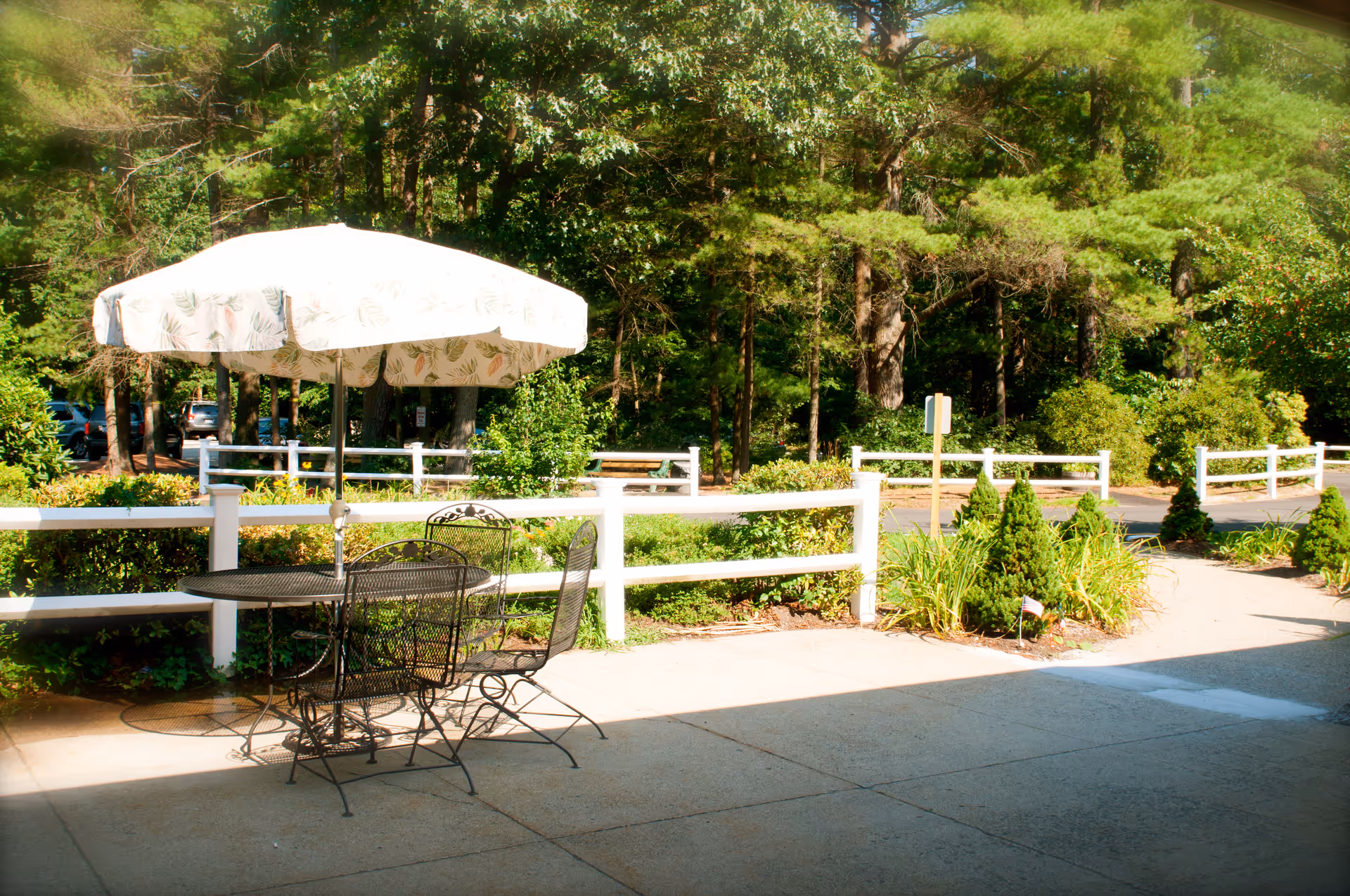Outdoor patio area with a metal table and chairs under a large white umbrella with a leaf pattern. The patio is adjacent to a garden with green bushes and trees, and a white fence separates the patio from a parking area and wooded background.