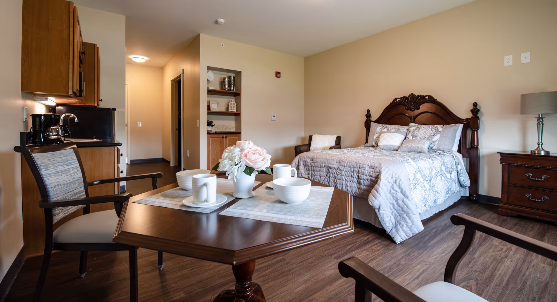 A cozy studio-style room with a made bed, bedside table, kitchenette, and a dining table set with cups and a vase of flowers.