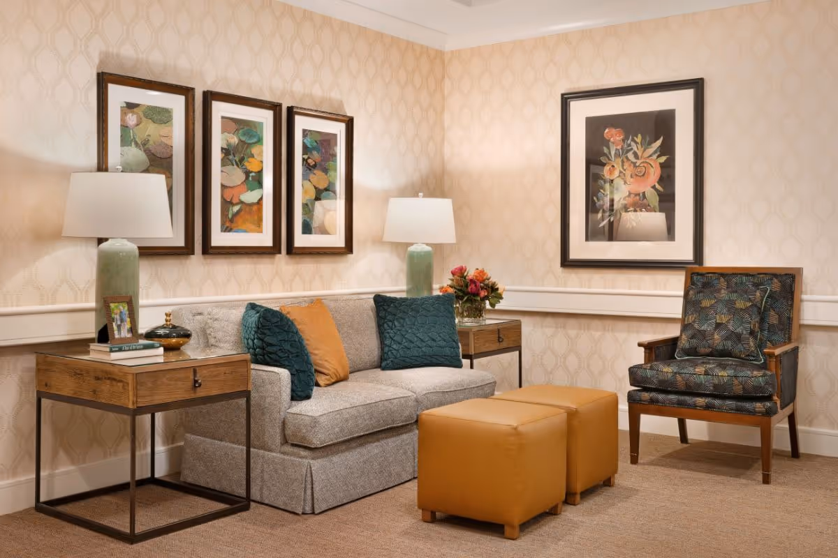 Comfortable seating area with a sofa, patterned armchair, two ottomans, side tables, lamps and framed artwork in a softly lit living room.