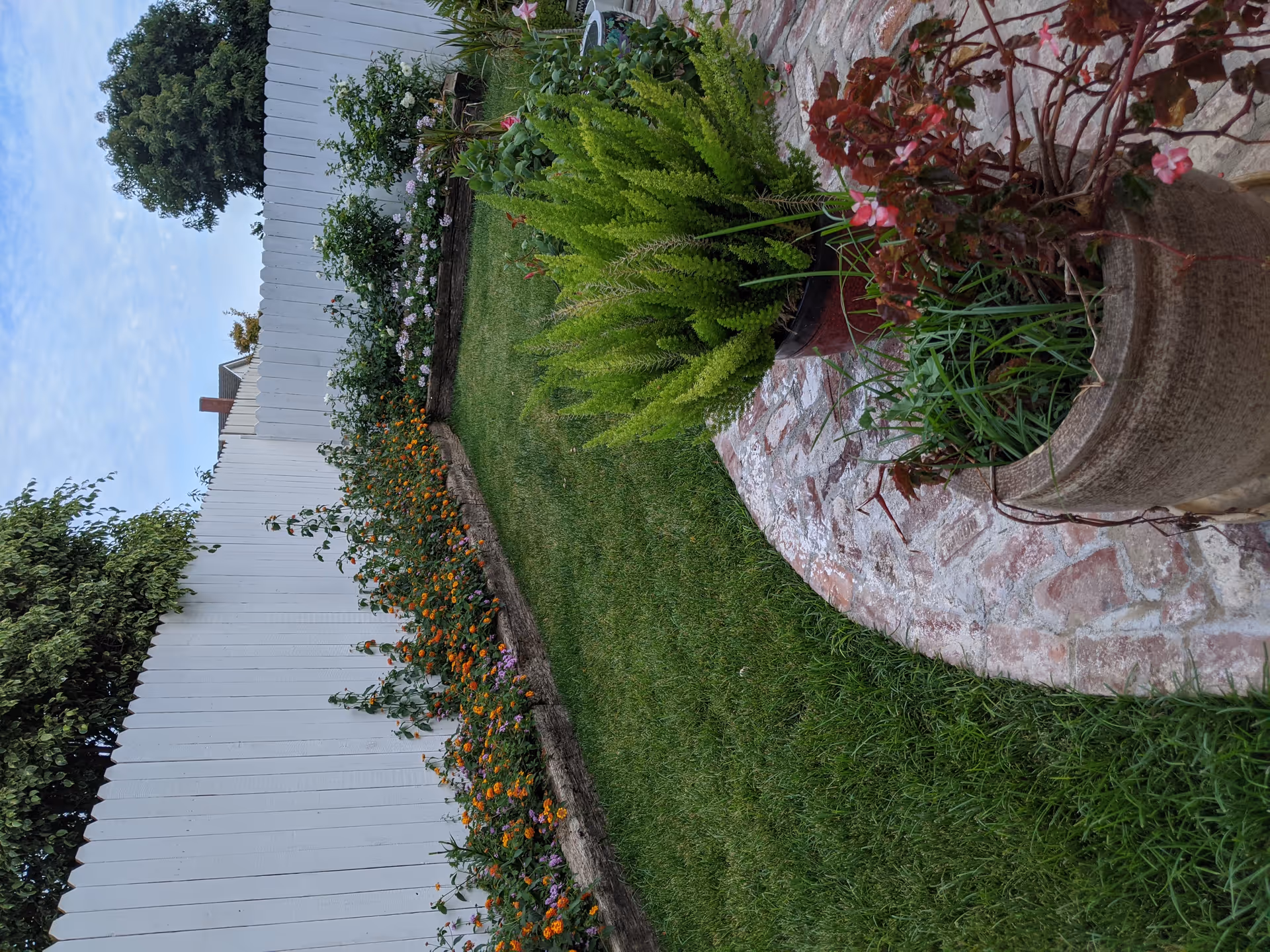 A well-maintained garden area with a white wooden fence in the background. There is a green lawn bordered by flower beds with orange and purple flowers. In the foreground, there are potted plants on a curved brick patio.