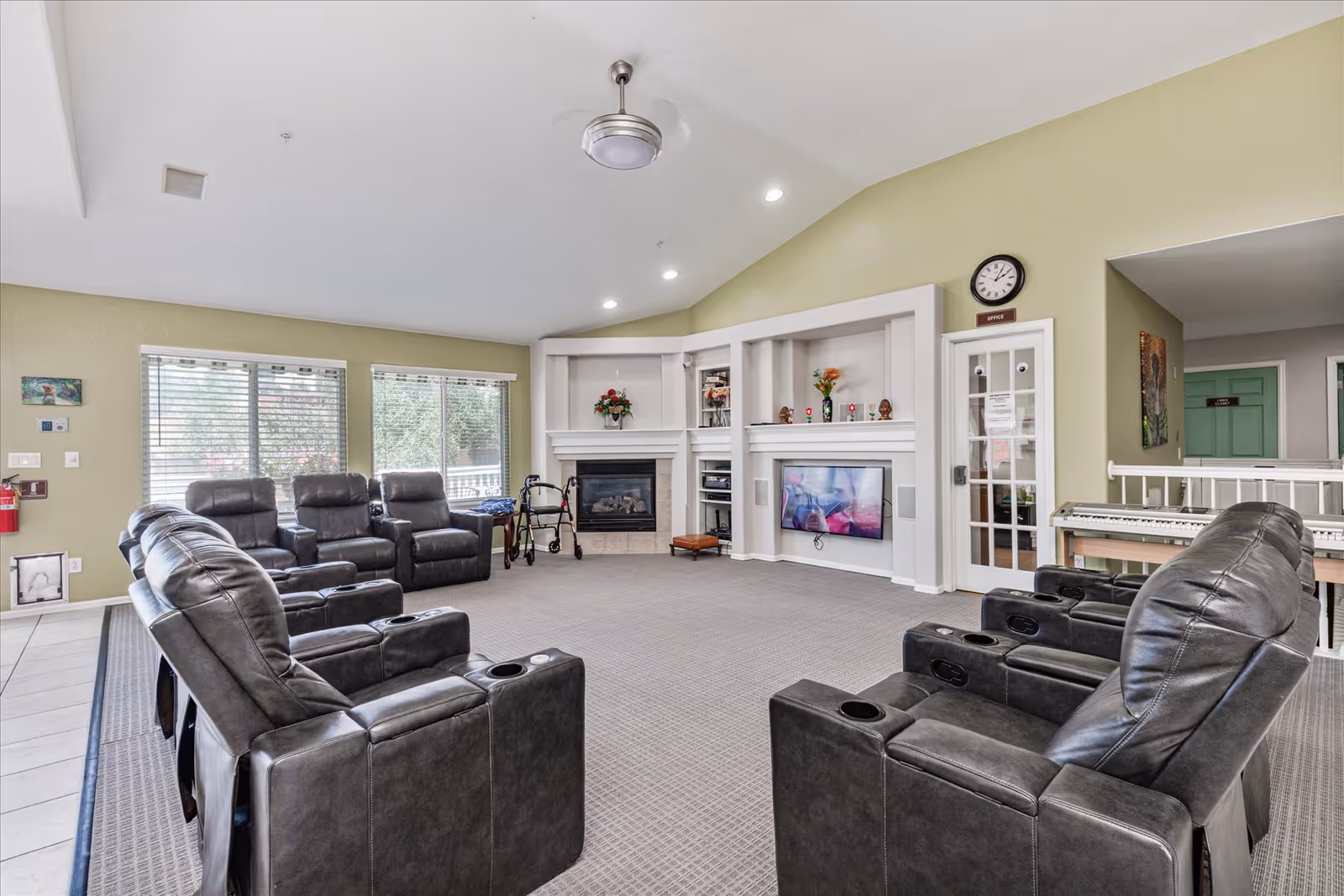 A spacious senior living facility common room with multiple black leather recliner chairs arranged in a semi-circle facing a wall-mounted TV and a fireplace. The room has light green walls, large windows with blinds, a ceiling fan, and a clock above a door labeled 'Office'. There is a walker near the fireplace and a keyboard on a stand to the right.
