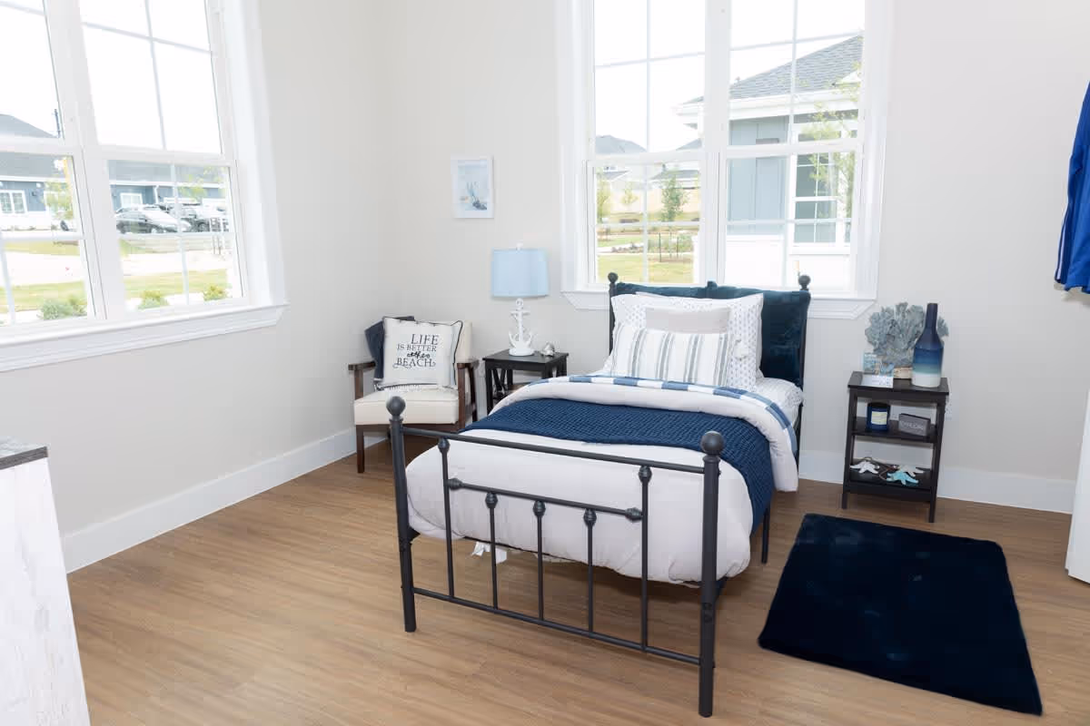A bright bedroom with a single black metal bed frame, neatly made with white and blue bedding. There is a small black nightstand with decorative items and a lamp, a chair with a cushion that reads 'Life is better at the beach,' and large windows letting in natural light. The floor is wooden with a dark blue rug beside the bed.