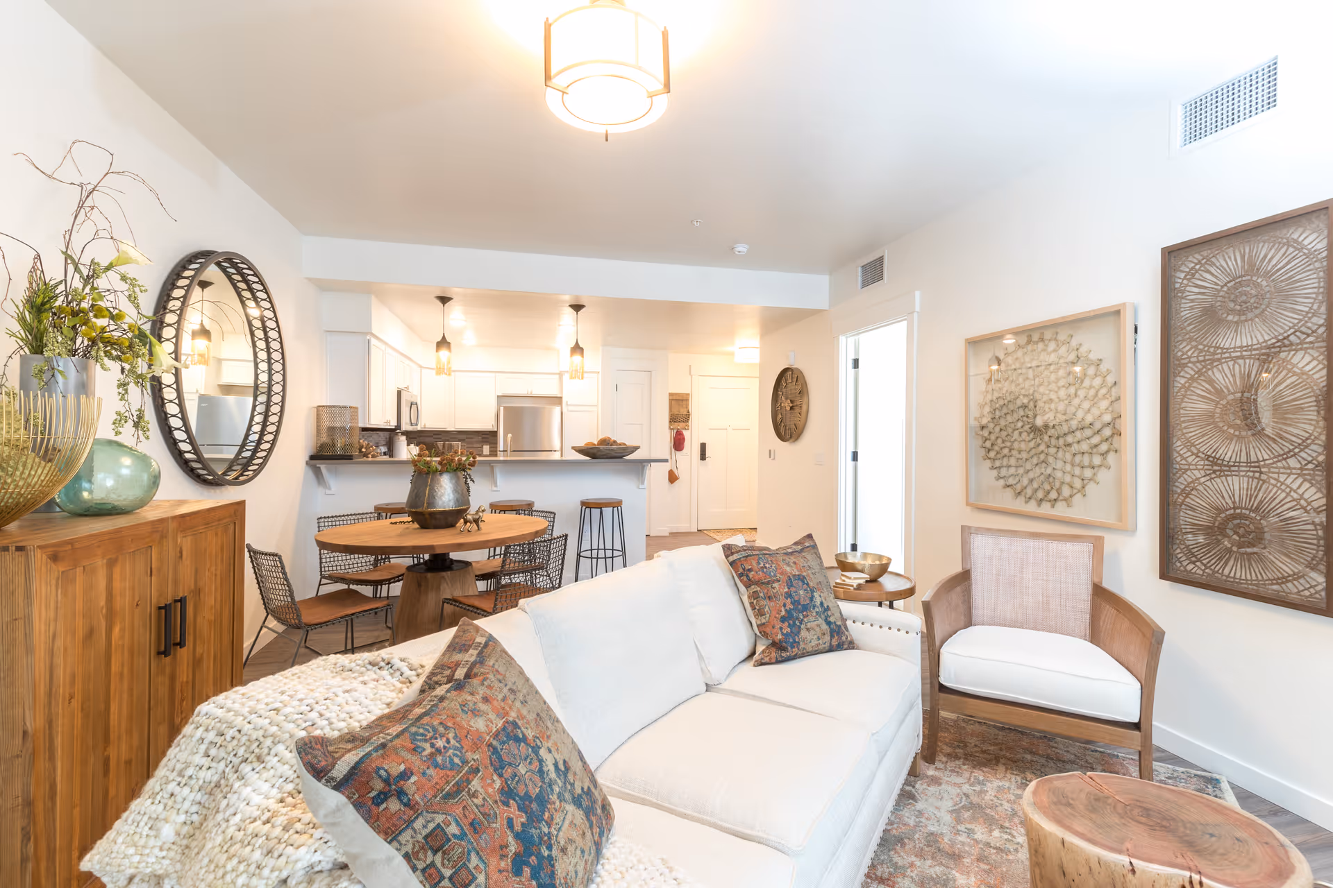 Bright and cozy living room area with a white sofa adorned with patterned pillows and a knitted throw blanket. A wooden armchair with a white cushion sits next to the sofa. The walls feature decorative art pieces. In the background, there is a dining area with a round wooden table and metal chairs, and a kitchen with white cabinets, stainless steel appliances, and pendant lights hanging above the counter. A wooden sideboard with plants and decorative items is on the left side of the image.
