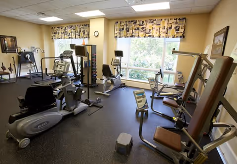 A well-lit exercise room with various fitness equipment including stationary bikes, weight machines, and a step platform. The room has large windows with patterned valances allowing natural light to enter, and framed artwork on the walls.